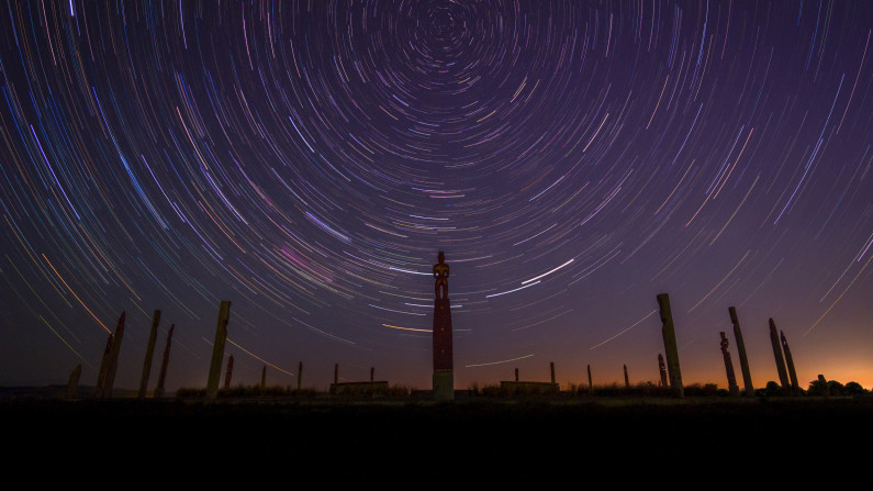 Star-streaks form a circular pattern above a circle of pouwhenua (carved pillars).