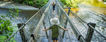 Two small children are crossing a wire swing bridge barefoot. The bridge is above a wide, slow-moving river. Native trees and a building are visible in the background.