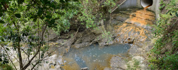 A culvert embedded in a hillside releases water into a small stream. The water has stained the concrete below the culvert an orange colour.