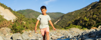 A boy is walking barefoot through a small bouldery river. Regenerating forest covers the hills behind him.