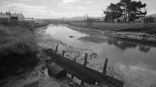A black and white photo showing a wide polluted stream with a metal barrel and partition in the foreground.