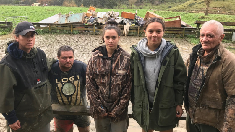 Five people stand in front of a muddy field and broken furniture piled up behind a fence.