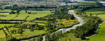 An aerial view of a river winding through a flat landscape. There are with green fields and trees beside the river and an urban area in the background.