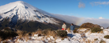 Two trampers sit on rocks in the snow and tussocks, enjoying the view of snow-clad Mt Taranaki.