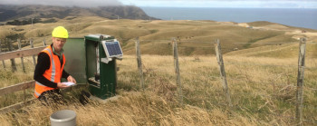 Inside a farm fence, a man wearing a hard hat and high-vis vest looks at a rain gauge. There is a solar panel beside him, and brown grass on the hills behind.