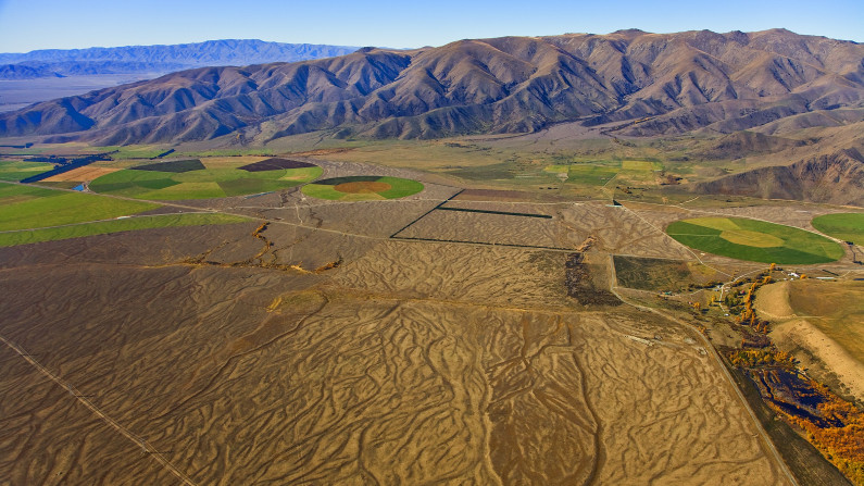 An aerial view with bare brown land in the foreground, green circles of irrigated land, and a range of hills behind.