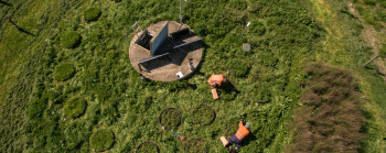 Aerial view of two people in high-vis vests kneeling down and looking at circles of darker green on a field.