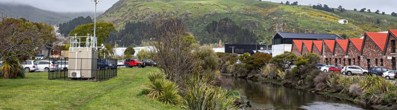 Photo of an air quality monitoring station, a small building with many sensors and aerials on the roof. It is located on a patch of grass next to a river, with industrial buildings and hills in the background.