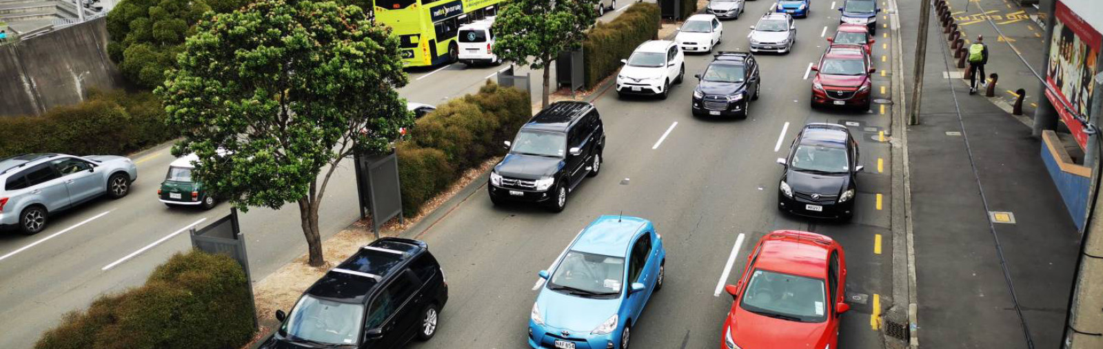 Photo of many cars stuck in slow moving traffic, in central Wellington. There are many tall buildings surrounding the busy six lane road, and some trees adjacent to the road.
