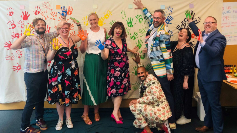 Eight people excitedly stading in front a banner painted with hand-prints in rainbow colour order (red, orange, yellow, green, blue, purple).