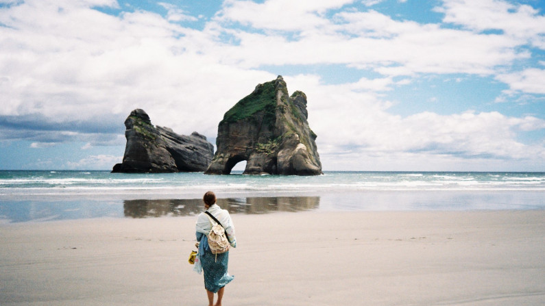 A landscape of a beach with rock formations sticking out of the ocean. In the foreground is the back of a woman standing on the sand, looking out to sea.
