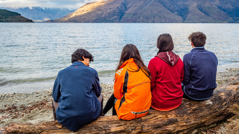 Four people sitting on a log on a lakeshore looking towards mountains on the other side.