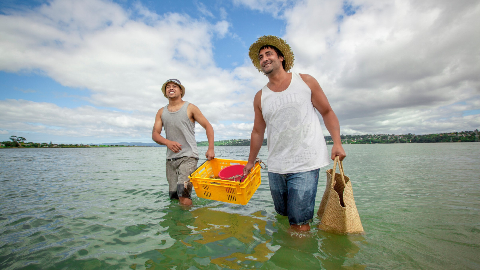 Two men smiling, one carrying a woven bag and both carrying a basket together as they wade through a body of water.