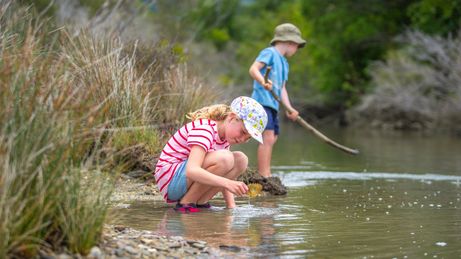 A young girl crouches near a river with a shell while a boy standing nearby with a stick examines the river shore.