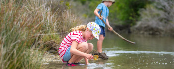 A young girl crouches near a river with a shell while a boy standing nearby with a stick examines the river shore.
