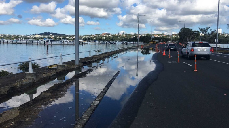 One lane of a road beside the sea is covered in water, and road cones are in place to divert traffic.