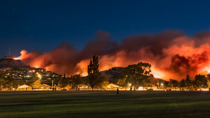 A large wildfire at night, with red smoke above the hills.