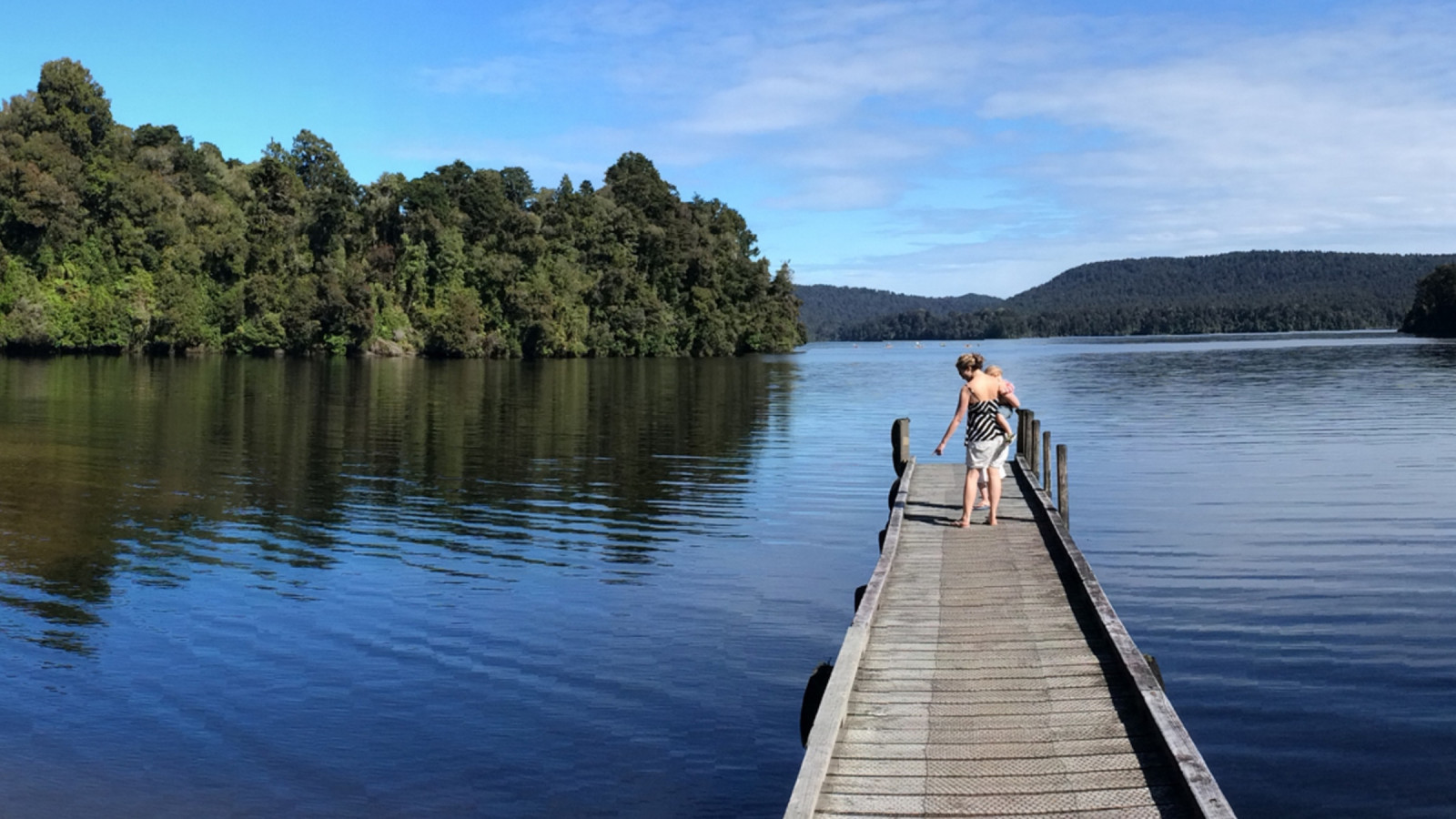 A person is walking along an old-fashioned jetty that extends into a lake. Native forest cloaks the hills behind and the sky and water are clear and blue.