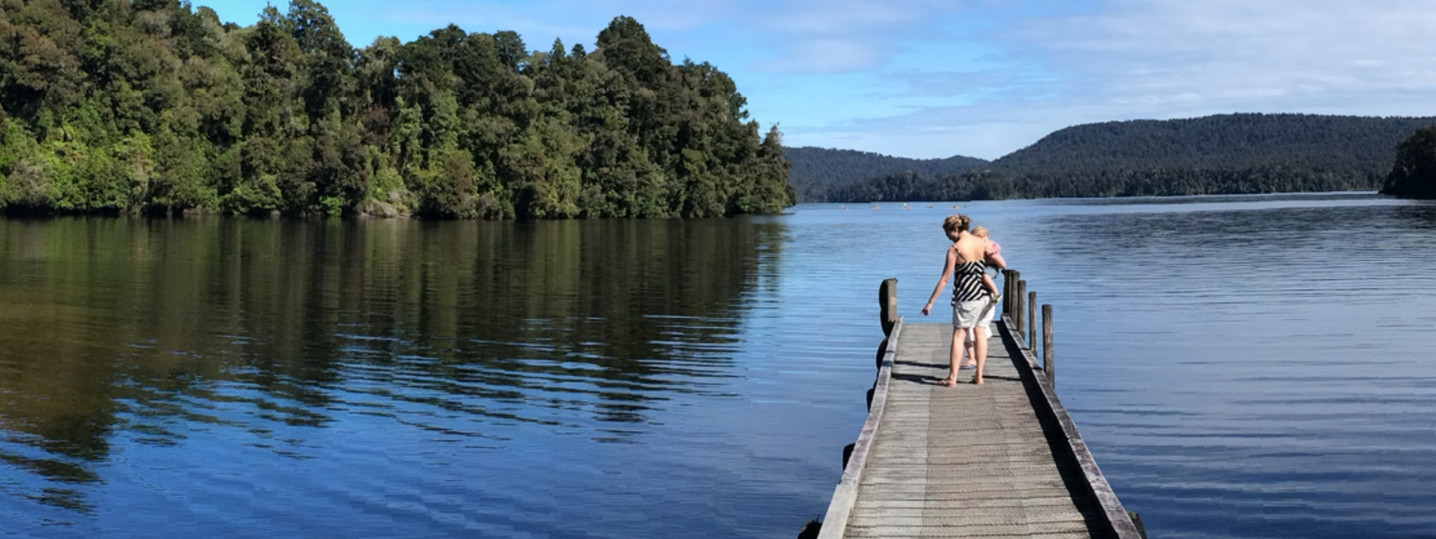 A person is walking along an old-fashioned jetty that extends into a lake. Native forest cloaks the hills behind and the sky and water are clear and blue.