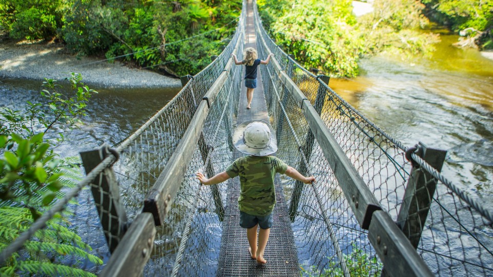 Two small children are crossing a wire swing bridge barefoot. The bridge is above a wide, slow-moving river. Native trees and a building are visible in the background.