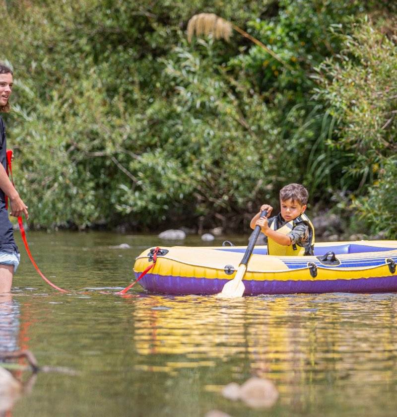 An older kid standing in a river, helping a young kid who is sitting in an inflated rate with a paddle.