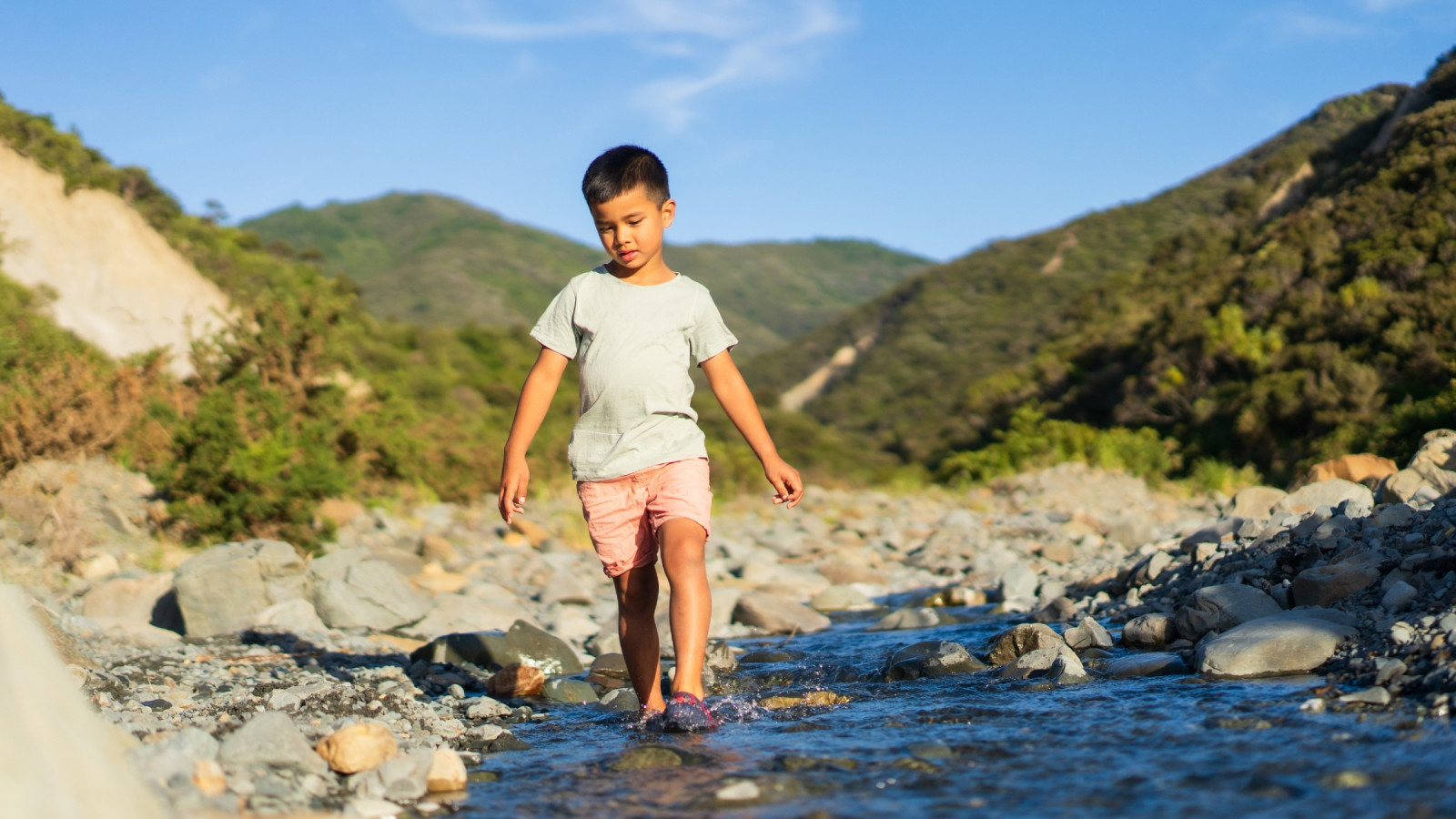 A boy is walking barefoot through a small bouldery river. Regenerating forest covers the hills behind him.