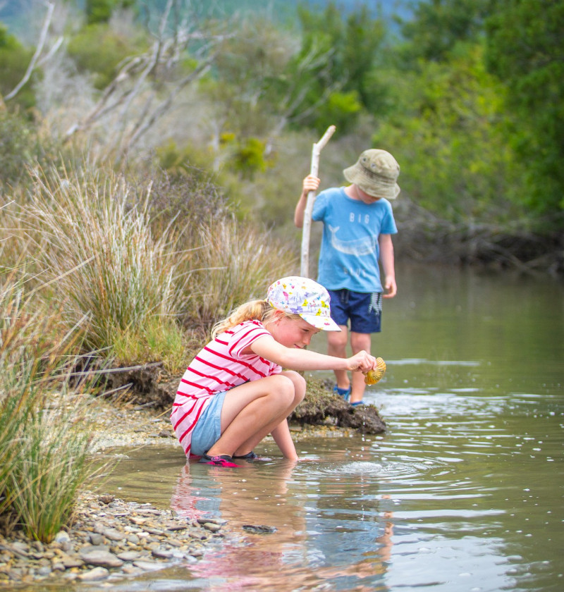 A young girl crouches near a river with a shell while a boy standing nearby with a stick examines the river shore.