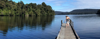 A person is walking along an old-fashioned jetty that extends into a lake. Native forest cloaks the hills behind and the sky and water are clear and blue.