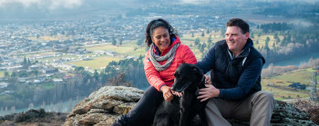 Photo of two people with a black dog, sitting on a rocky outcrop, overlooking a small Central Otago town. There is a river and low clouds surrounding the town.