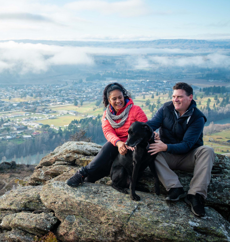 Photo of two people with a black dog, sitting on a rocky outcrop, overlooking a small Central Otago town. There is a river and low clouds surrounding the town.