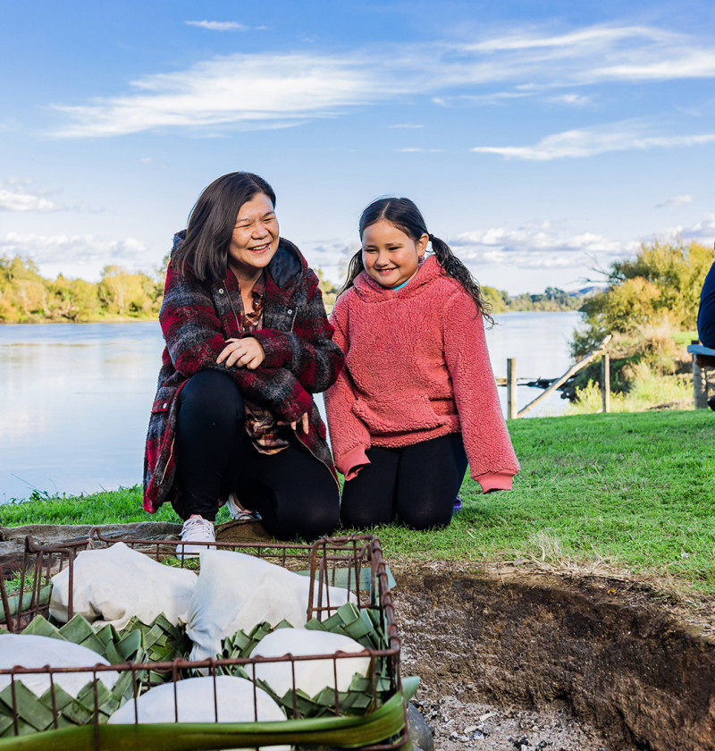 An adult and child smile whilst watching over a hāngī by the side of a river.
