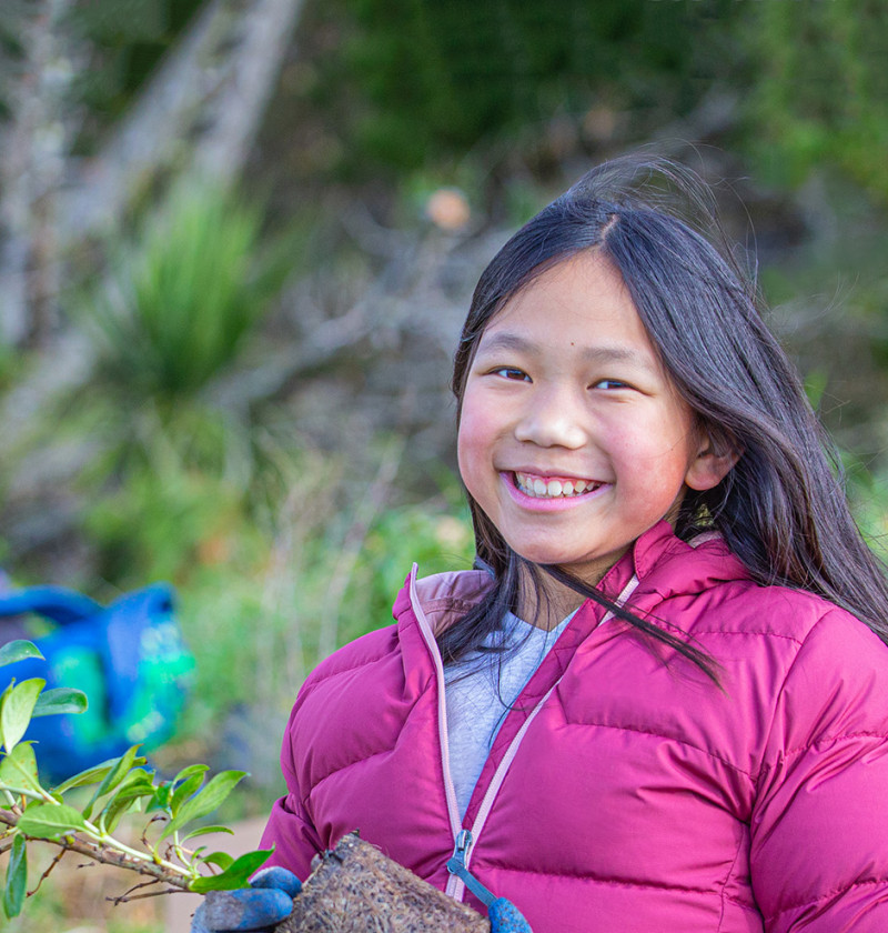 A young person smiling and holding a native plant at a tree-planting event.