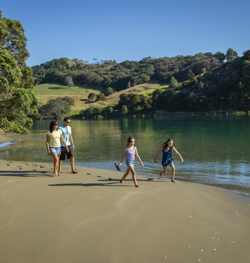 Two adults and two children walk along the sandy bank of a river.
