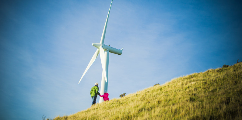 Two people stand by a wind turbine on a hill.