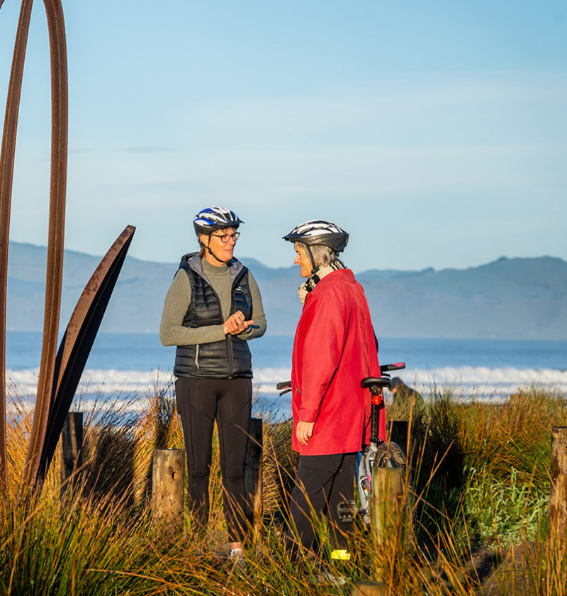 Two women talking next to a sculpture near the seaside. 