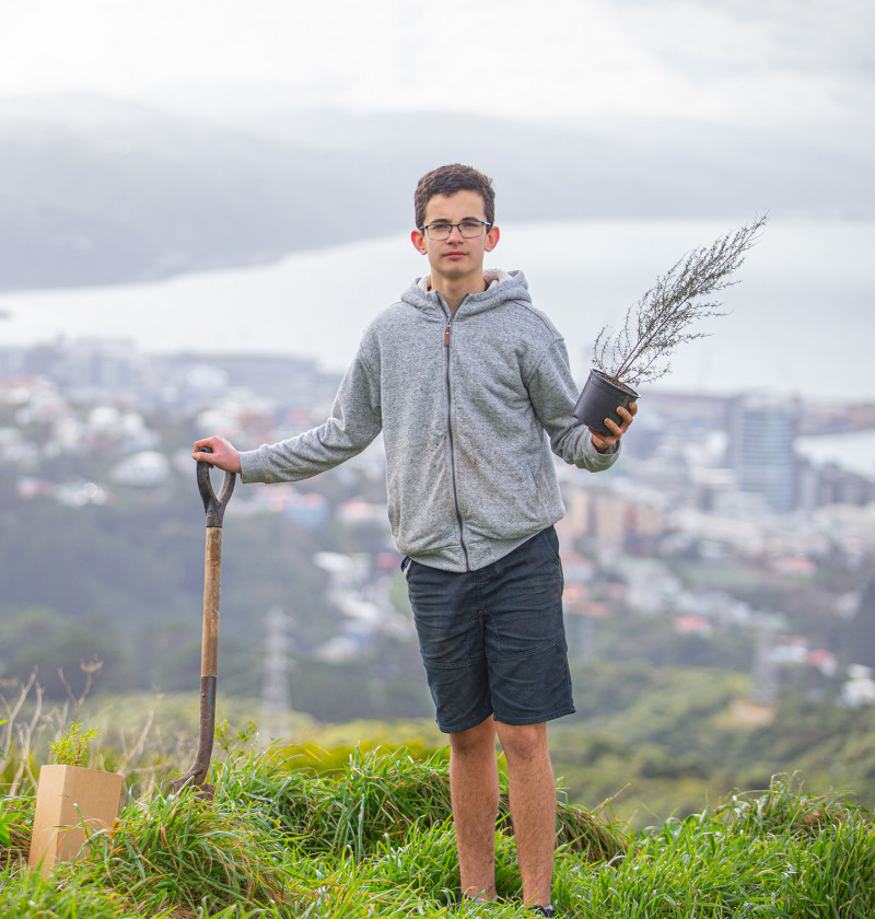 A teenage boy leaning on a shovel and holding a native tree. A city can be seen behind him.  