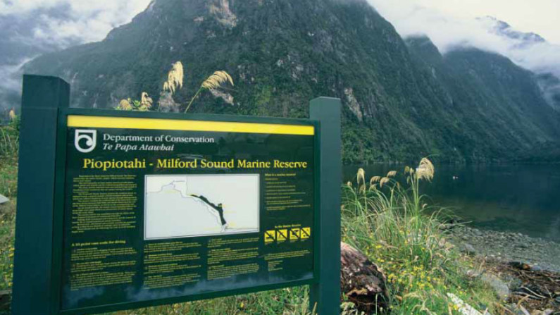 A sign reading 'Piopiotahi - Milford Sound Marine Reserve', with mountains and water behind it.