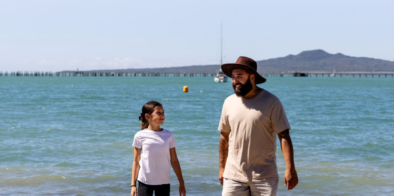 A girl and a man standing on the shore of a beach. Rangitoto in the background