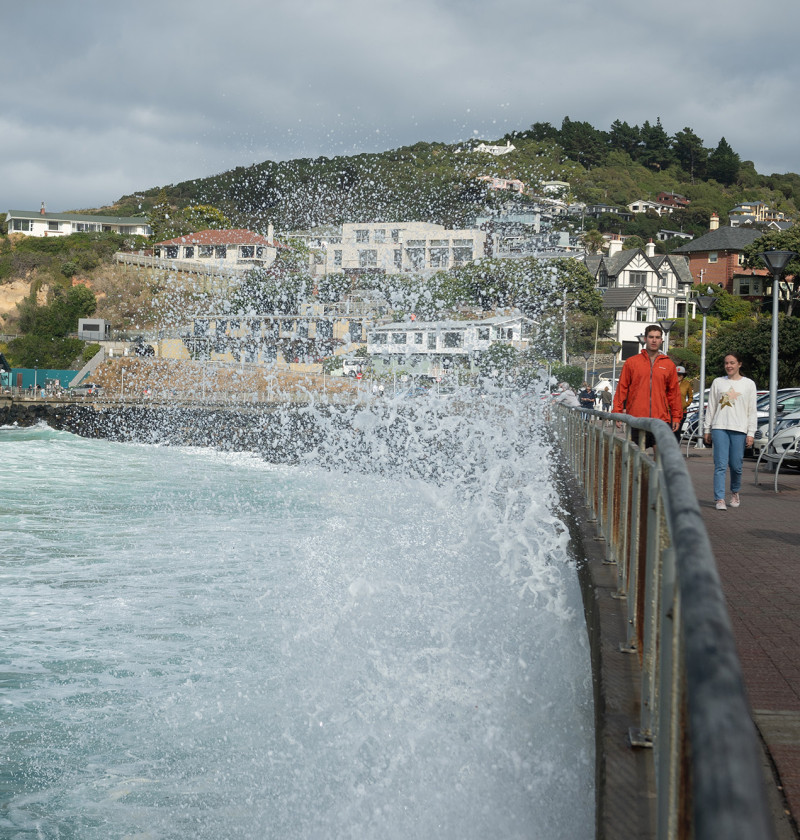 Two people walking alongside a waterfront, with big waves splashing up on them. Houses on hills are in the background.