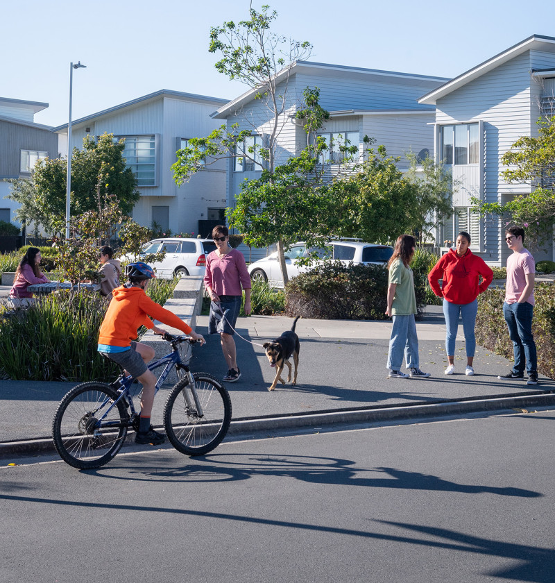 Medium-density housing with various groups of people outside. They are talking, seated at a table, riding a bike and waking a dog.
