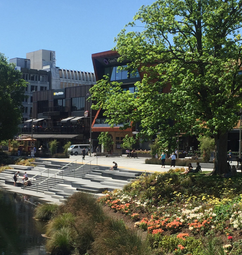A photo showing people interacting with an urban environment in Christchurch. There are steps leading to the river and trees and other plants growing. 