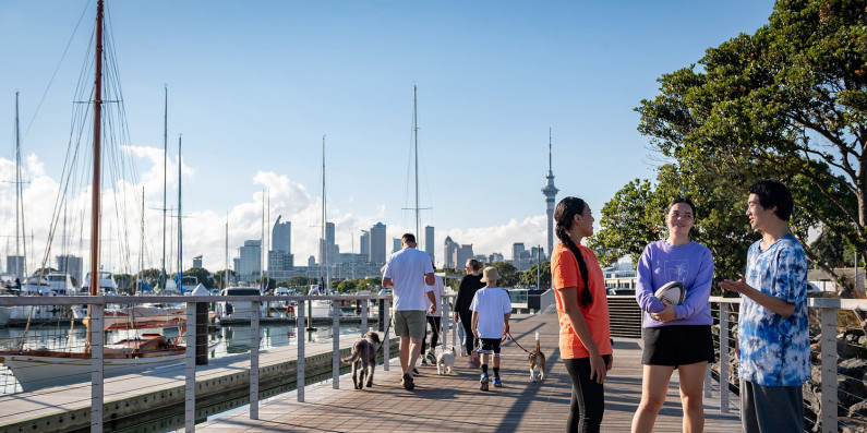 In the foreground, three young adults stand on a walkway, chatting. In the mid-ground, a family are walking their dogs. In the background are trees, Auckland's sky tower, buildings and the viaduct.