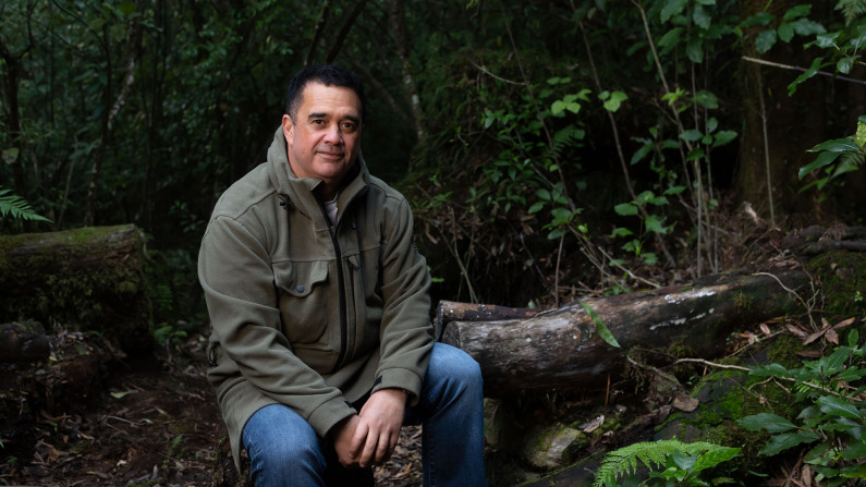 Professor Rangi Matamua (Chief Advisor Matariki and Mātauranga Māori) sits on a log in a forest area.