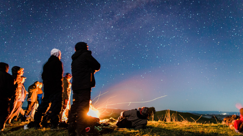 A group of people stand by a small fire on a hill and look up towards the clear sky filled with stars.