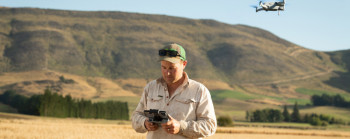 A farmer standing in a field holding a drone control. Farm hills can be seen in the background. 