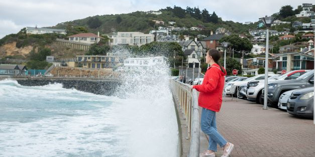 Girl standing on the street in front of cars as a wave from st clair beach Dunedin hits the retaining wall.