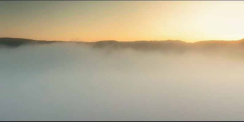 Photo of a hilly landscape covered by clouds, with the sun behind it.