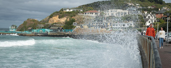 Two people walking alongside a waterfront, with big waves splashing up on them. Houses on hills are in the background.