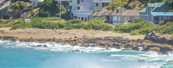 Houses by the beach, with green hills in the background and waves crashing on the rocks in the foreground.