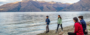 Two young adults are walking on the shore by some pristine water and two other young adults are sitting on a log on the shore. Mountains are in the background.